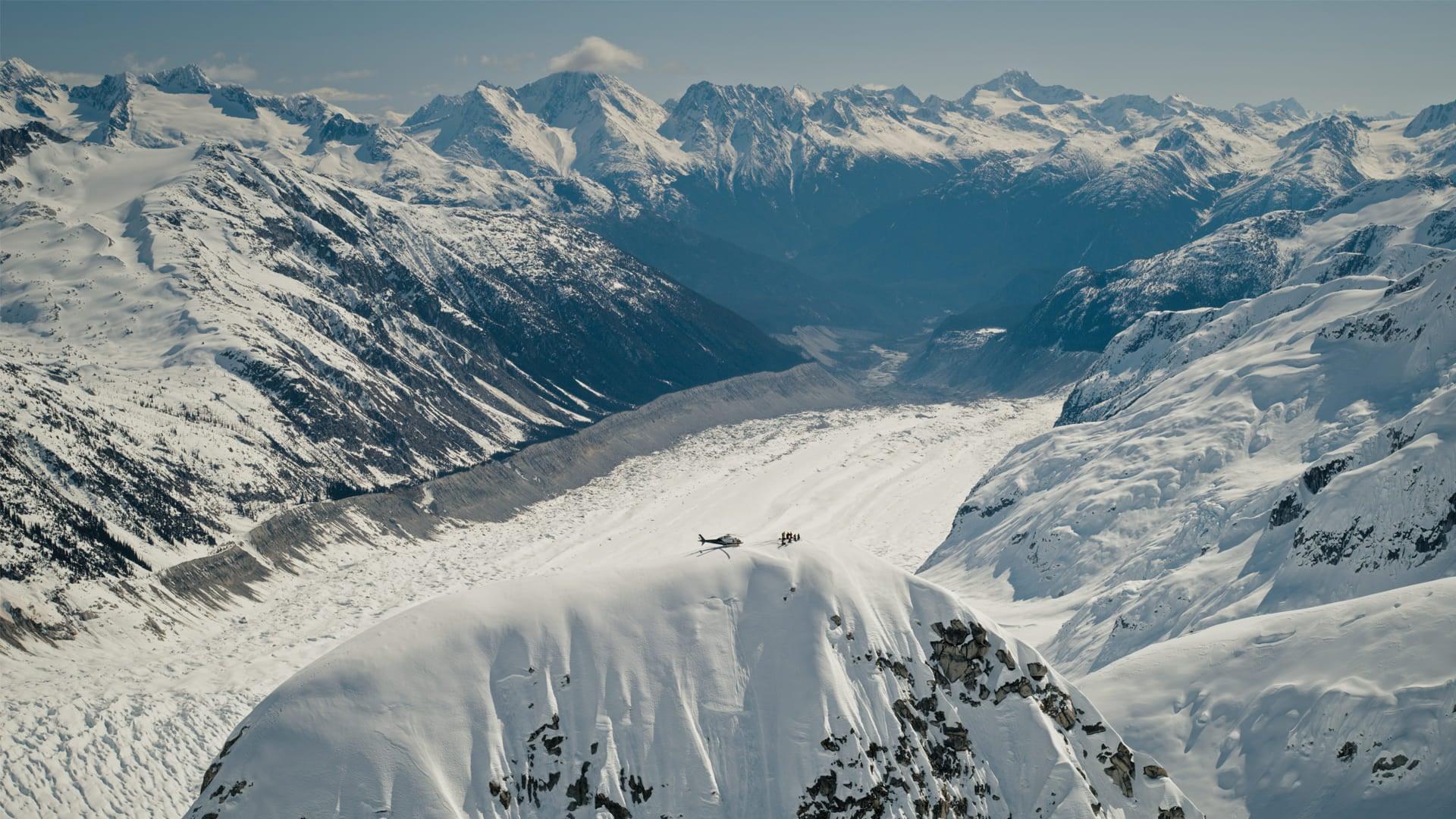 Helicopter and small group standing on a snowy mountain ridge above a vast glacier, surrounded by rugged, snow-covered peaks.