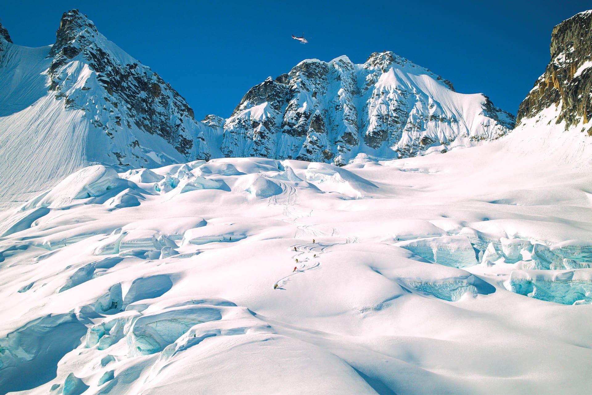 a group of five heli skiiers skiing down a glacier with a red, white, and blue helicopter flying overhead