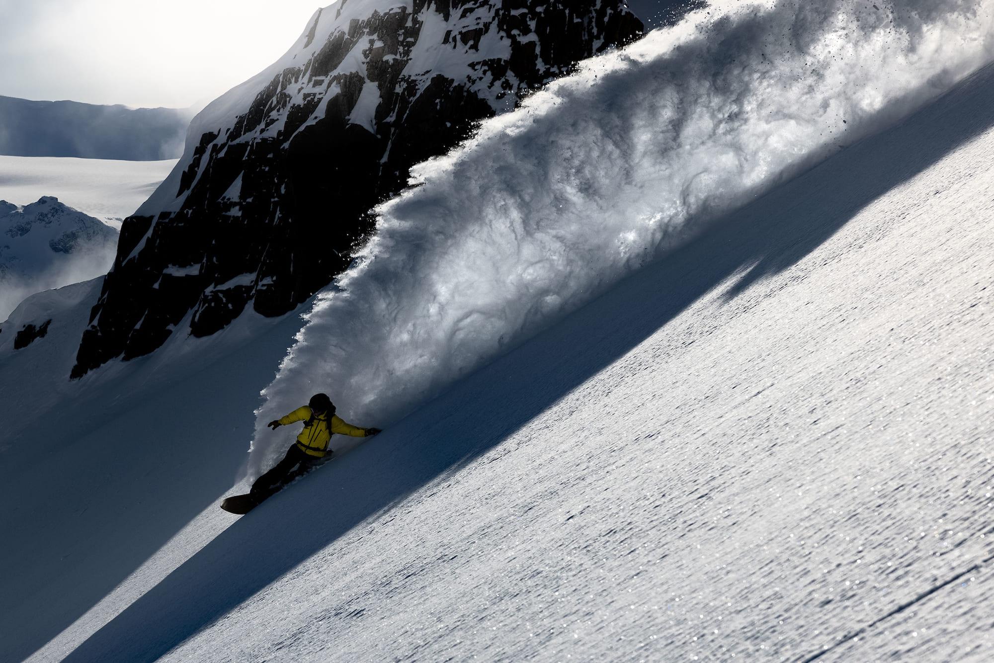 a snowboarder in a yellow jacket making a turn and spraying a big wall of snow
