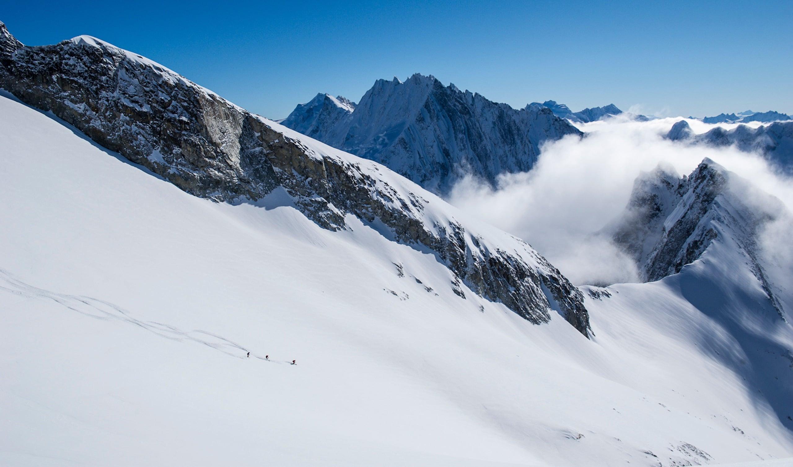 Two skiers traverse a snowy slope below jagged mountain peaks emerging from clouds.