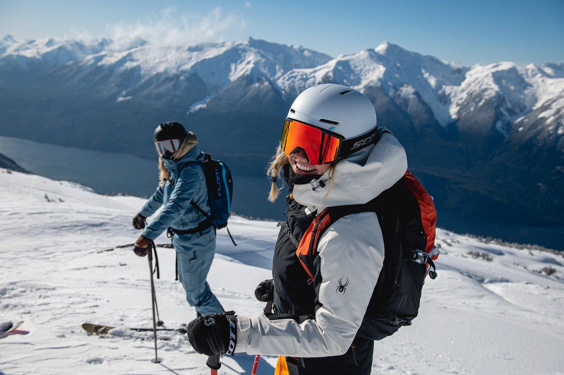 Two skiers taking a rest at the top of a mountain. The skier in the foreground is smiling